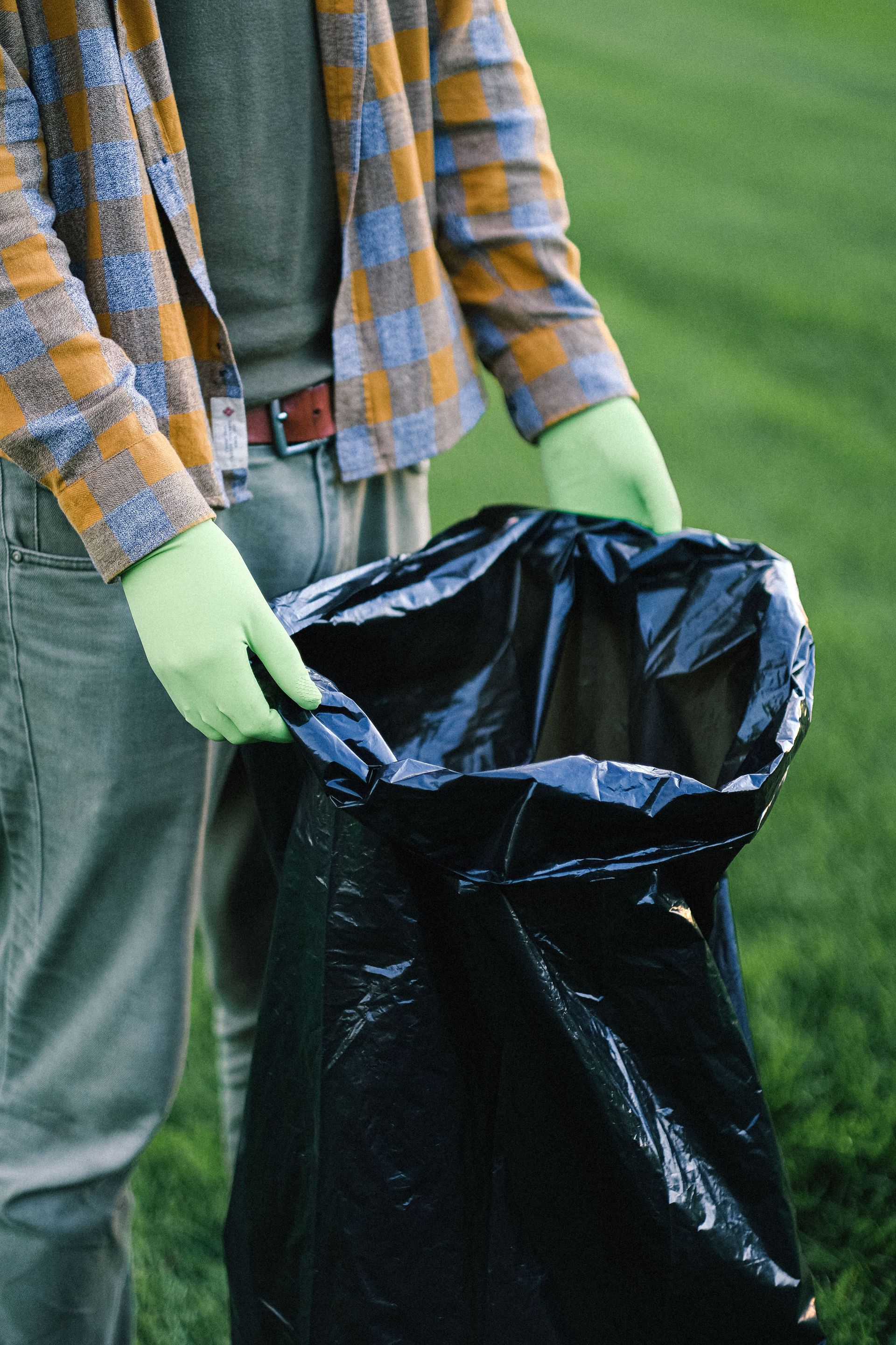 Person wearing gloves holding a trash bag in a green field, emphasizing volunteer cleanup efforts.
